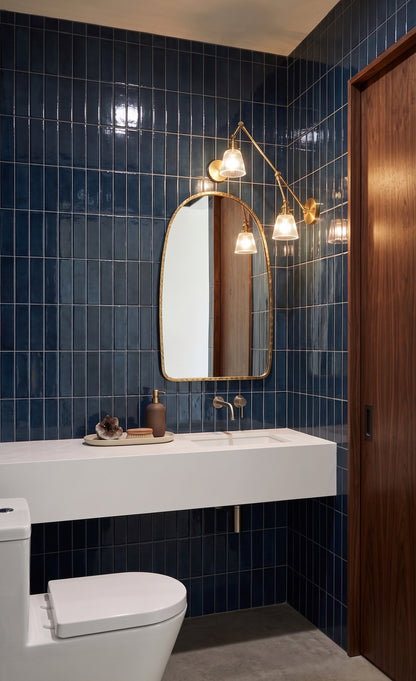 A modern bathroom interior with deep blue glossy wall tiles, a white floating vanity, a white toilet, a gold-framed oval mirror, and a wall-mounted brass sconce with three glowing bulbs.