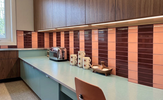 A modern kitchen counter area with dark wood cabinets, a mint green countertop, and a glossy tiled backsplash in alternating vertical stripes of terracotta orange and deep burgundy. A chrome toaster, patterned ceramic mugs, and a small wooden bowl sit on the counter, with a brown chair in the foreground.
