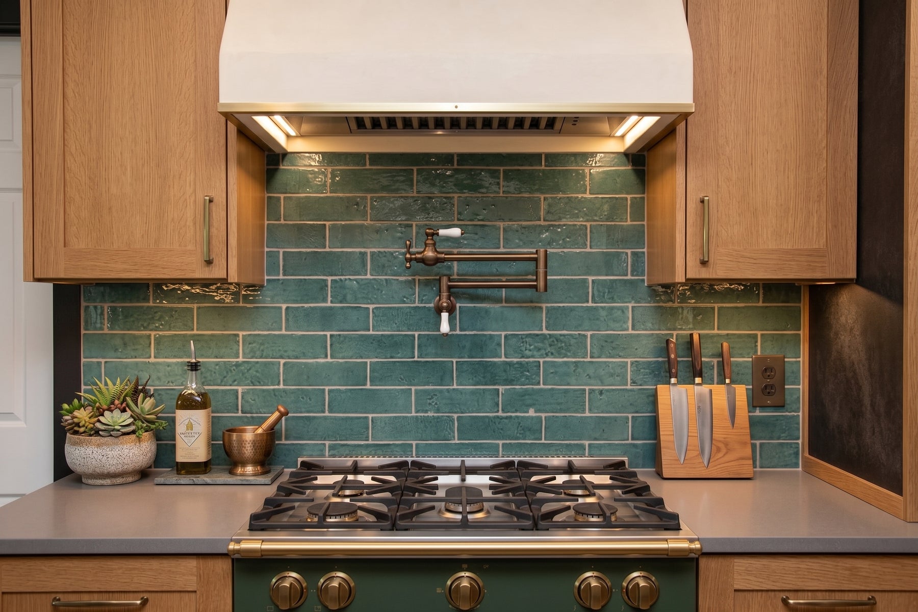 A kitchen backsplash made of glossy teal-green subway tiles behind a gas stove, with light wood cabinets, a white range hood, and a gray countertop.