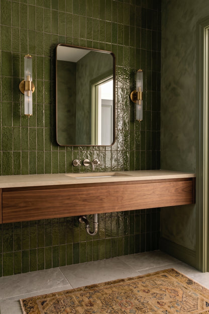 A modern bathroom vanity featuring a floating wooden sink with a light stone countertop, set against a glossy green tiled wall with a rectangular mirror and two brass wall sconces.