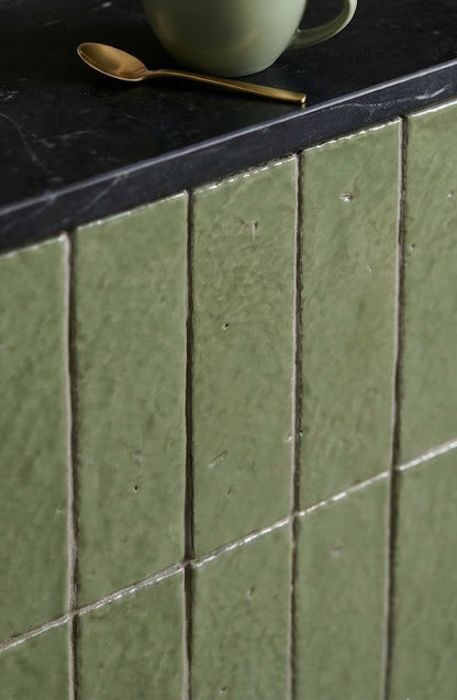 Moss green glazed thin brick by Clay Imports with a rustic, uneven finish, installed vertically below a dark countertop, styled with a gold spoon.