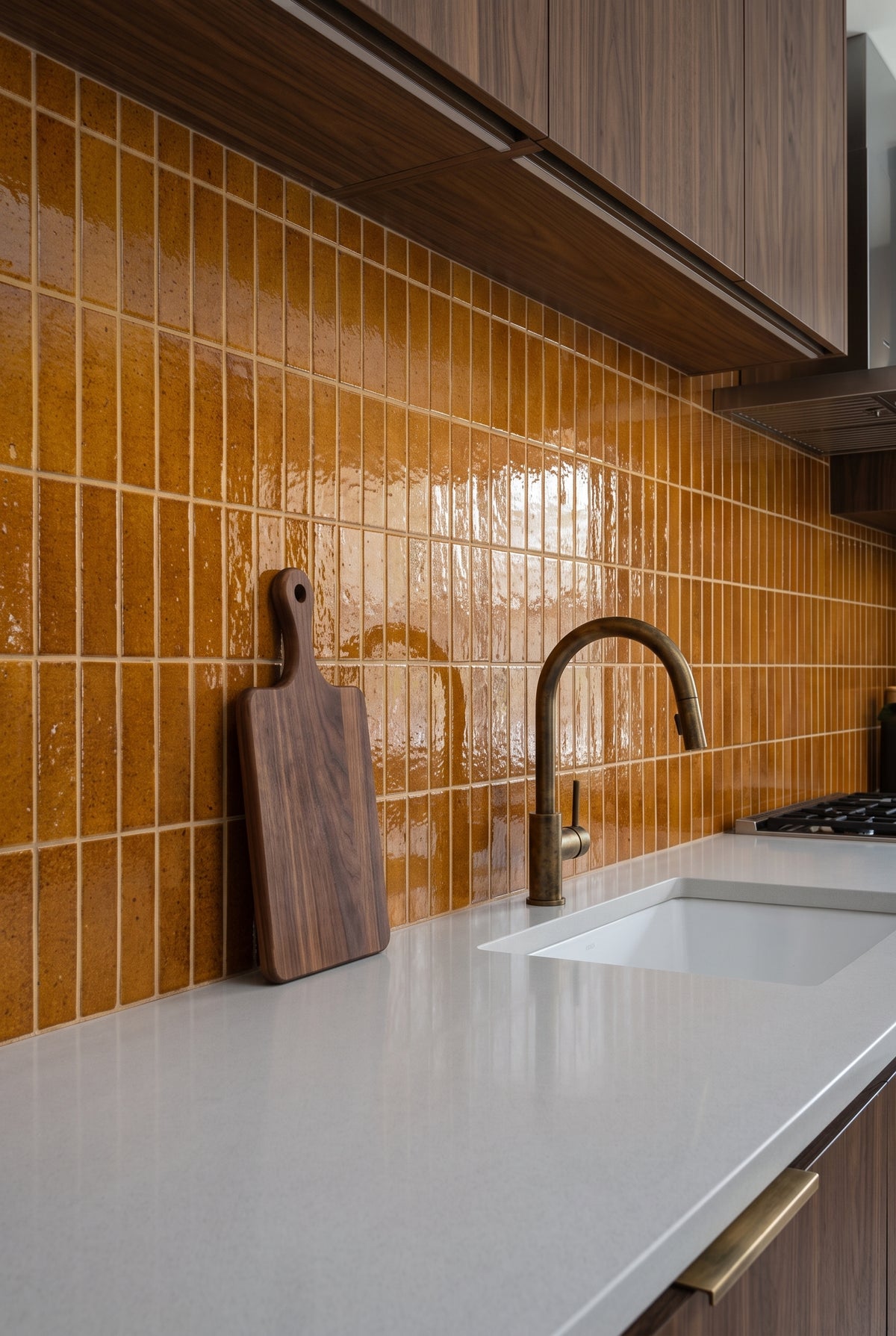 A wooden cutting board rests against glossy mustard yellow subway tiles in a modern kitchen with a white quartz countertop.