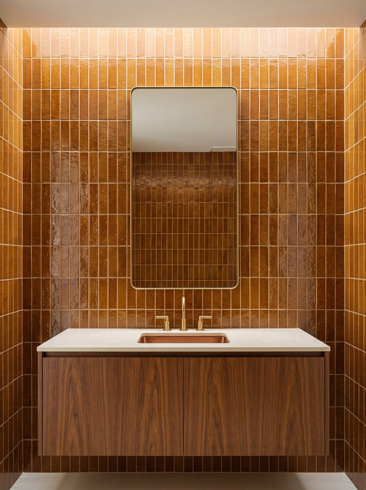 A modern bathroom vanity featuring a white countertop and a wooden cabinet sits against walls covered in glossy brown zellige tiles beneath a brass-framed mirror.