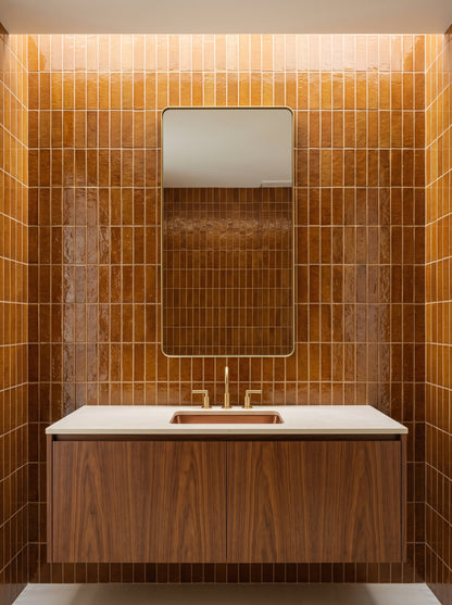 A modern bathroom vanity featuring a white countertop and a wooden cabinet sits against walls covered in glossy brown zellige tiles beneath a brass-framed mirror.