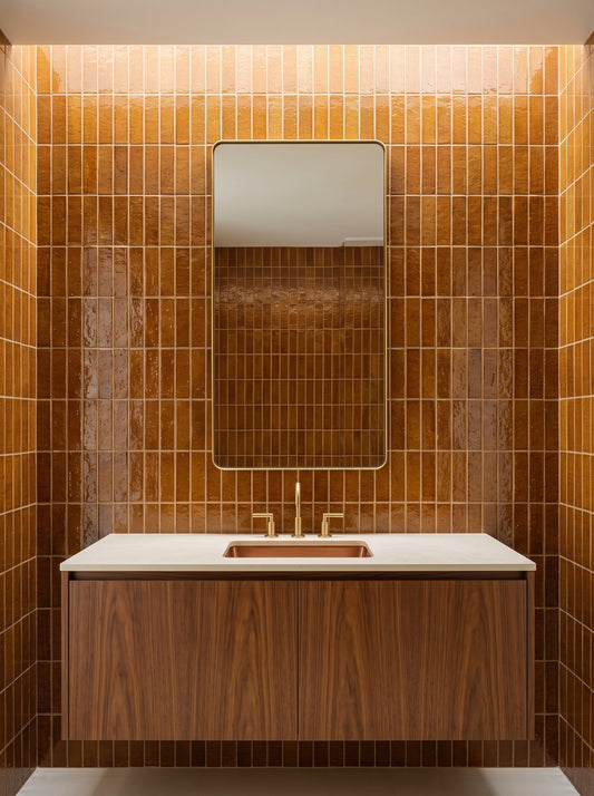 A modern bathroom vanity featuring a white countertop and a wooden cabinet sits against walls covered in glossy brown zellige tiles beneath a brass-framed mirror.