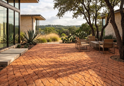 A terracotta-colored interlocking patio paver system arranged in a geometric diamond pattern, forming a spacious outdoor patio in front of a modern house with glass walls, trees, and outdoor seating.
