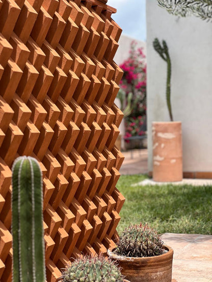 A terracotta-colored geometric outdoor wall panel with a repeating 3D starburst pattern, shown in a garden setting with cacti and grass.