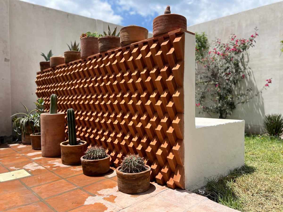 A terracotta-colored geometric wall planter made of interlocking wedge-like blocks, shown outdoors in a courtyard with potted cacti and succulents, white walls, grass, and a blue sky.