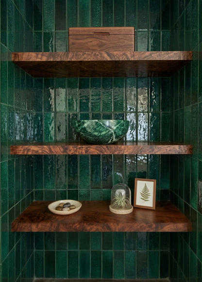 Three rich walnut wooden shelves styled with decor sit against a striking backdrop of glossy emerald green vertical ceramic tiles.