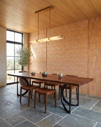 A modern dining room scene featuring a rectangular wooden dining table with a dark wood top and sculptural black metal bases, surrounded by matching wooden chairs. The room has a distinctive herringbone-patterned brick wall, a wood-paneled ceiling, and a stone-tiled floor, illuminated by a long linear pendant light.