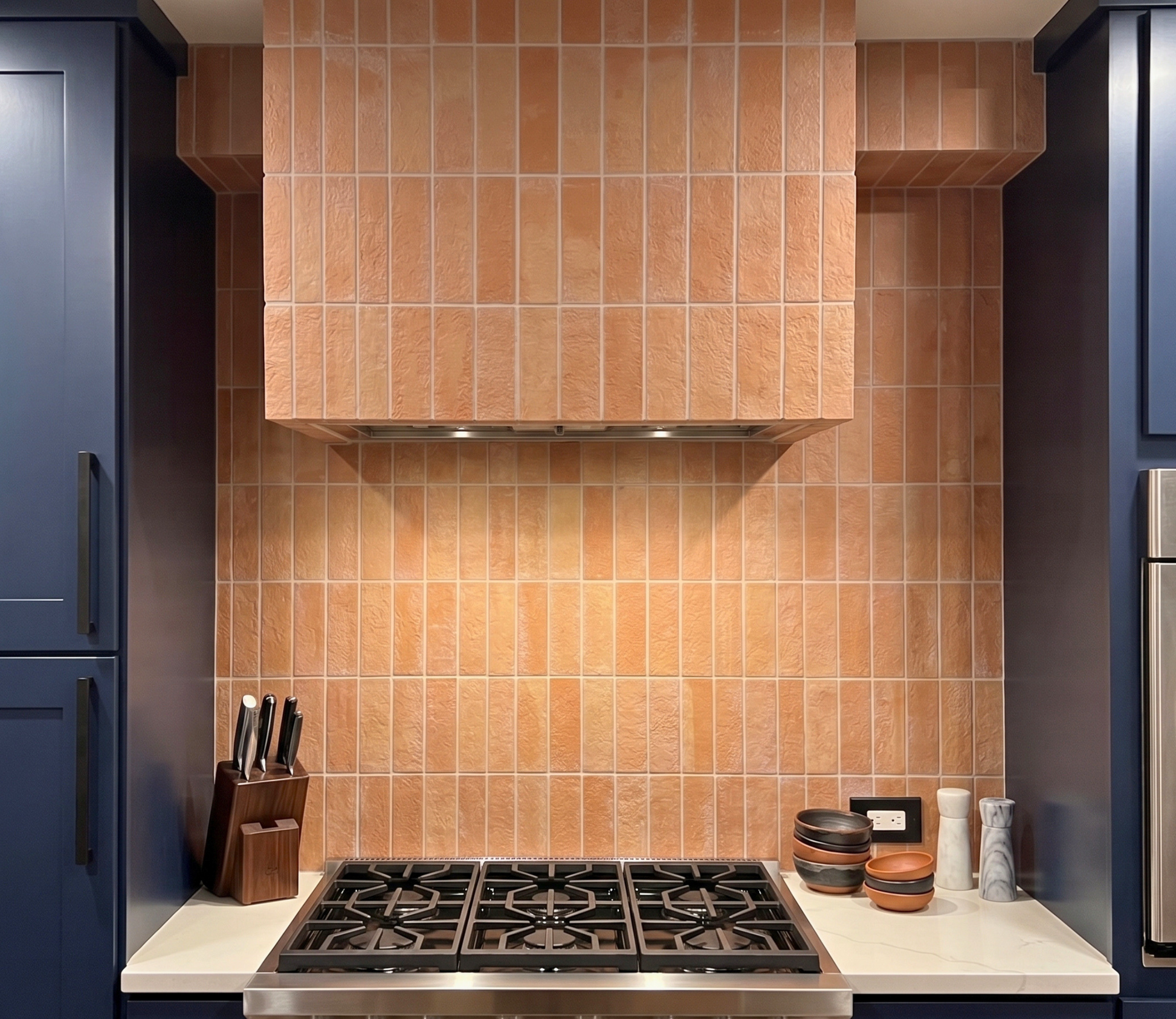 A modern kitchen backsplash featuring rectangular terracotta orange tiles on the wall and range hood surround, paired with dark navy blue cabinets, a white countertop, and a stainless steel gas range.
