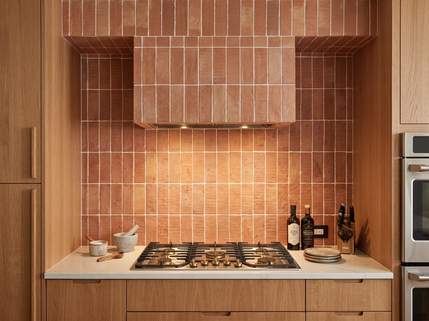 A modern kitchen backsplash featuring textured terracotta-colored rectangular tiles arranged in a grid pattern, installed behind a gas cooktop and a matching range hood. The surrounding cabinetry is made of light oak wood, and kitchen items like wine bottles, a mortar and pestle, and plates are neatly arranged on the white countertop.