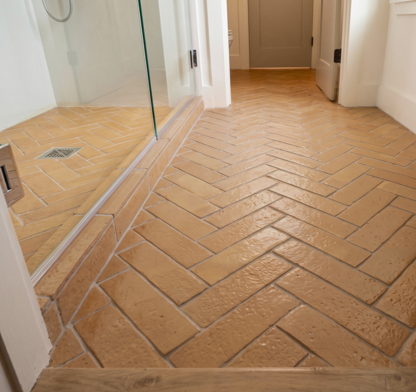 Terracotta-colored herringbone floor tiles installed in a bathroom hallway, with a glass shower door on the left and white walls and doors in the background.