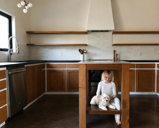 A child sits on a wooden kitchen island with a small white dog, set in a modern kitchen with wood cabinets, dark countertops, and a tiled backsplash.