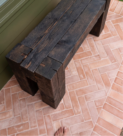 A rustic dark wood bench made from heavy timber blocks, positioned on a terracotta brick patio next to an olive green door.