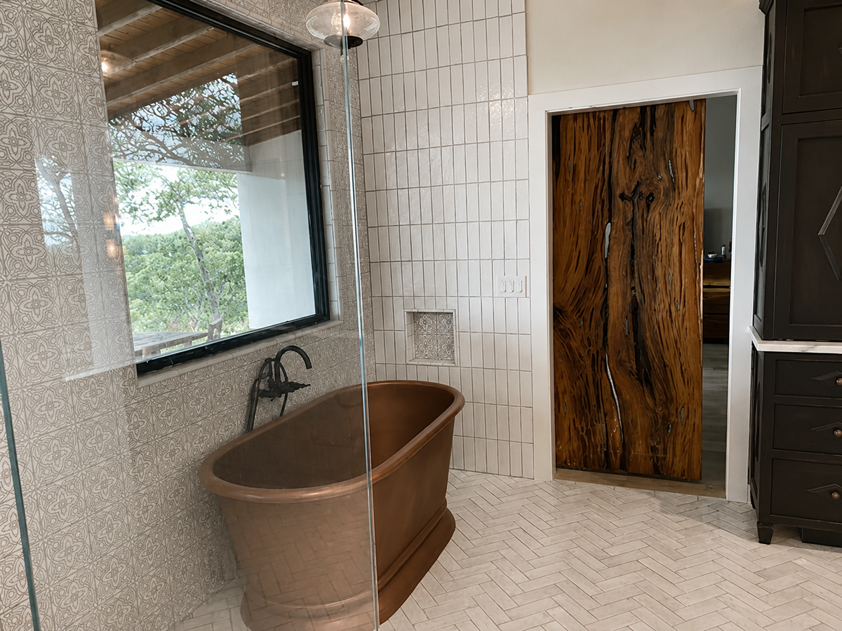 A freestanding copper clawfoot bathtub in a modern bathroom with white subway tile walls, a large black-framed window, and a dark wood door.