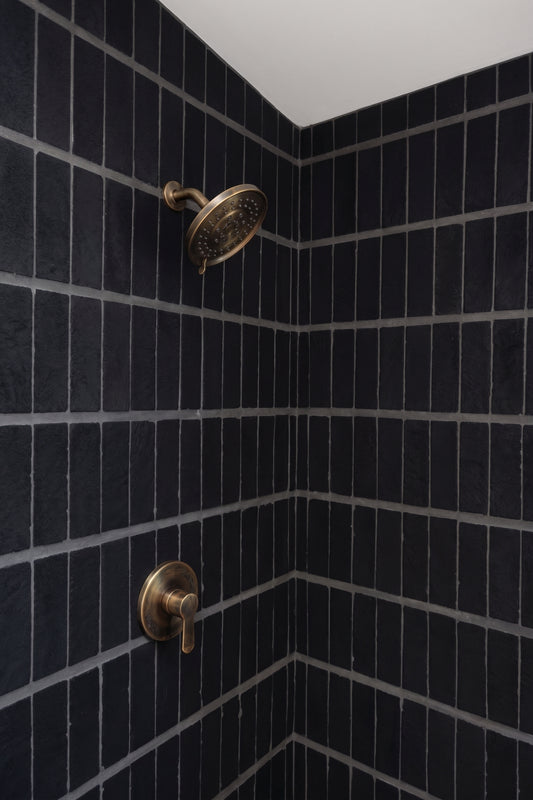 A corner shower stall with dark charcoal gray subway tiles and antique brass shower fixtures, including a round rain head and a single-handle valve.
