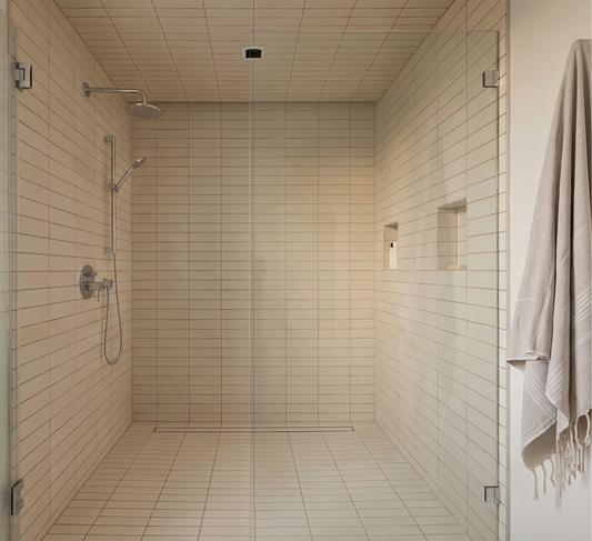 A modern walk-in shower room with beige rectangular wall and floor tiles, a glass door with metal hinges, a wall-mounted shower head and handheld shower, recessed niche details, and a folded cream towel hanging on the right side.