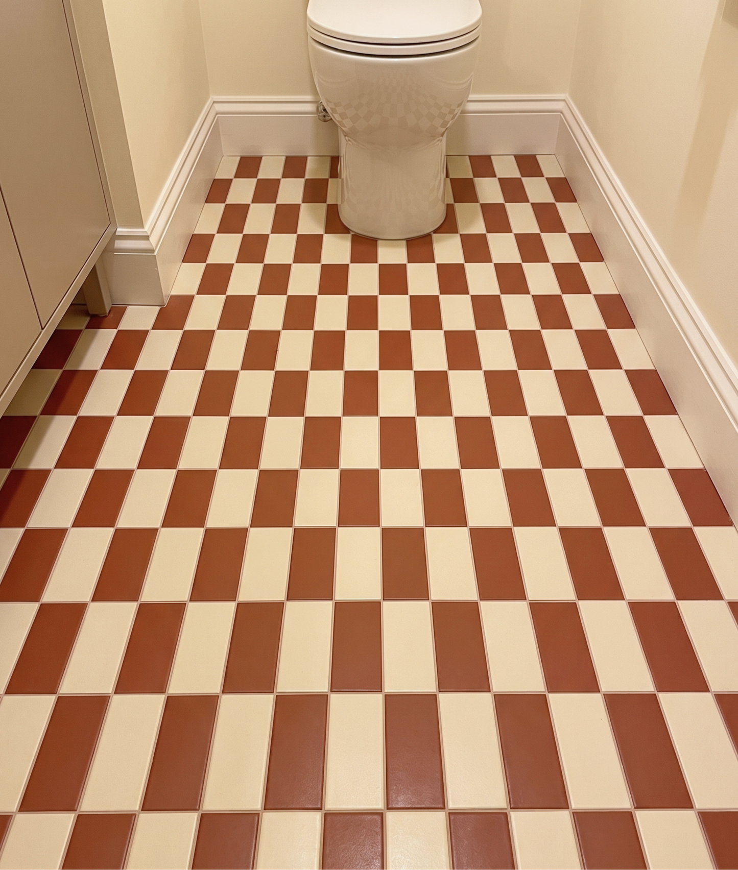 A checkered bathroom floor with alternating warm brown and cream square tiles, featuring a white toilet in the background against beige walls.