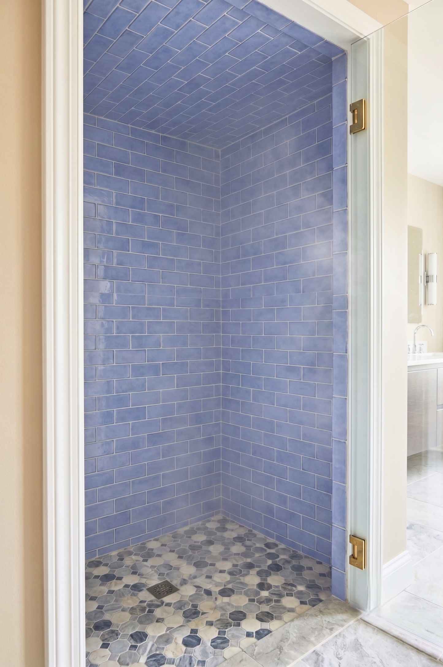 A walk-in shower stall with glossy light blue subway tile walls and a mosaic floor in blue, gray, and beige tones, shown from an adjacent room with cream walls and brass hinges.