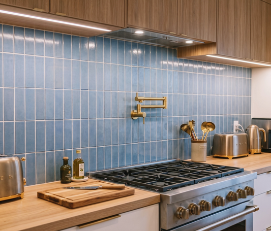 A modern kitchen scene with glossy light blue square wall tiles, light wood countertops, a stainless steel gas range, brass wall-mounted faucet, toaster, kettle, and cutting board.