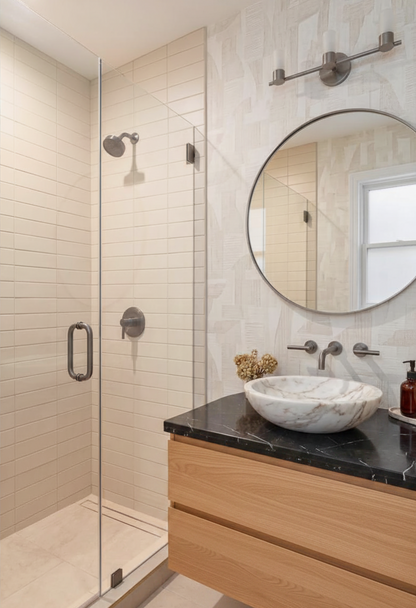 Modern bathroom vanity with a round mirror, wall-mounted brass light fixture, white marble vessel sink, black marble countertop, and light wood drawers, next to a glass-enclosed shower with beige tile walls.