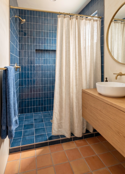 A modern bathroom interior featuring a walk-in shower with blue mosaic tile walls and floor, a cream linen shower curtain, a wooden floating vanity with a white vessel sink, and a round wall mirror.