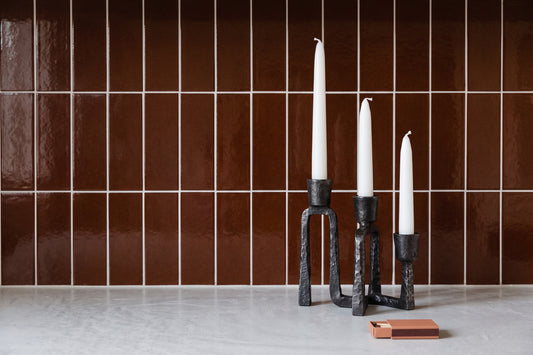 Three black metal candlestick holders with white taper candles, styled on a light gray surface against a glossy brown tiled wall.