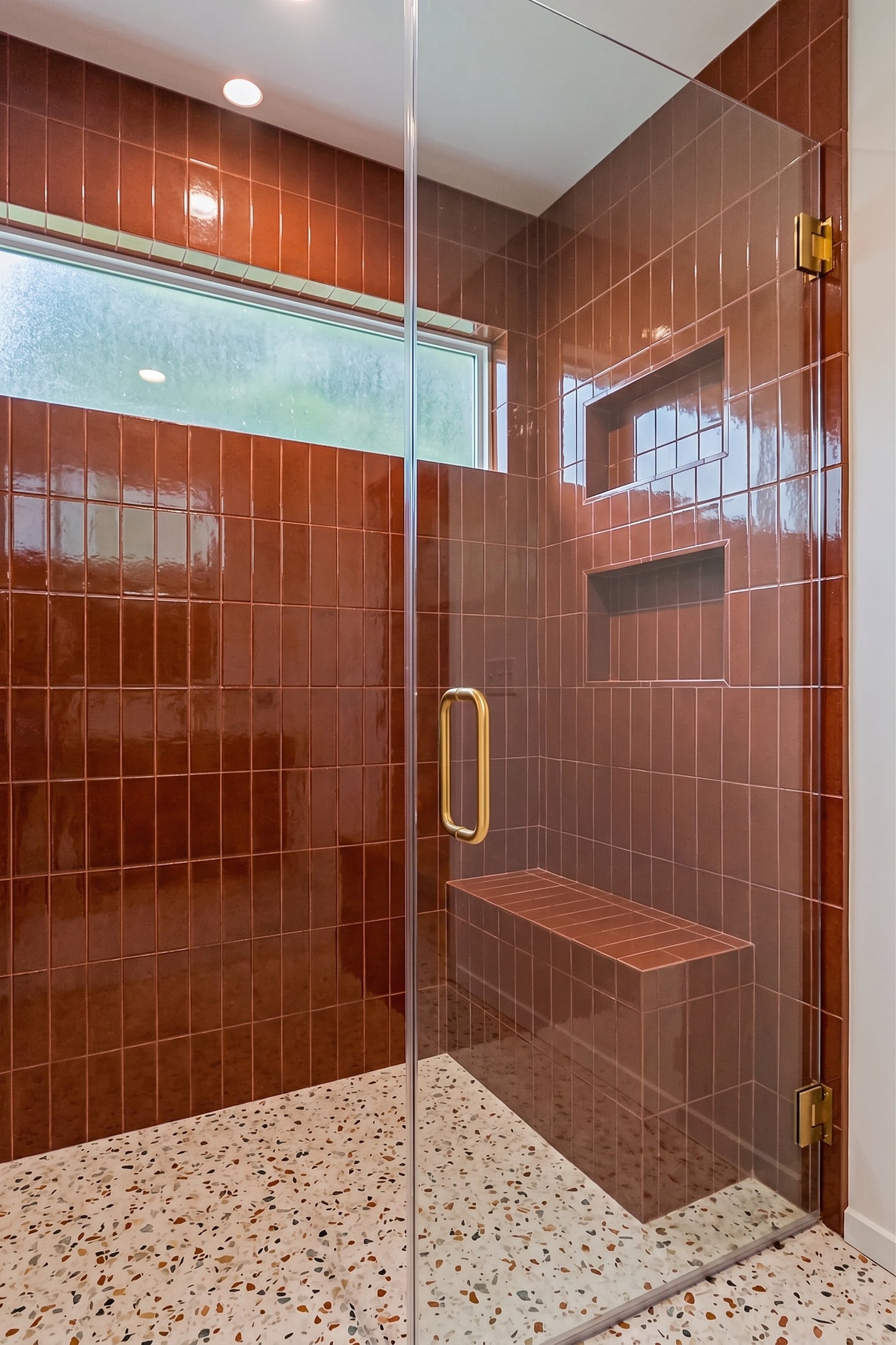 A modern walk-in shower enclosure with glossy reddish-brown wall tiles, a matching built-in bench, a terrazzo-style floor, and a clear glass door with a gold handle.