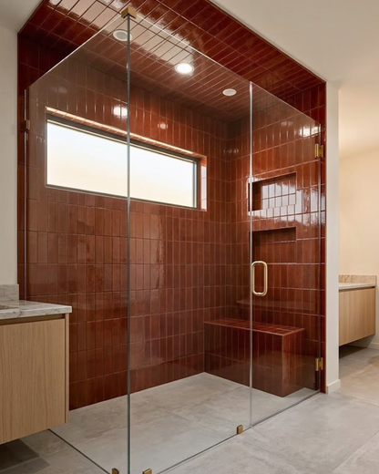 A modern glass shower enclosure with a walk-in shower featuring glossy reddish-brown tiled walls and floor, a built-in bench, and a small window, shown in a bathroom setting.