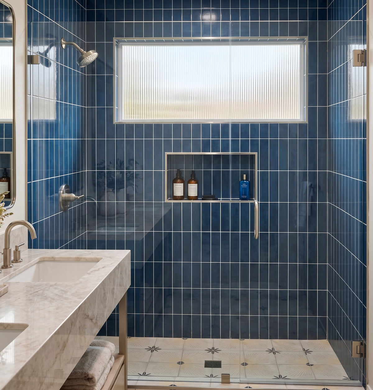A modern bathroom interior with glossy blue wall tiles, a freestanding marble vanity with a chrome faucet, a glass shower enclosure, and a recessed shelf holding two bottles and a blue container.