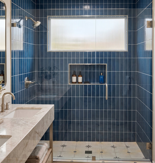 A modern bathroom interior with glossy blue wall tiles, a freestanding marble vanity with a chrome faucet, a glass shower enclosure, and a recessed shelf holding two bottles and a blue container.