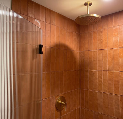 A modern shower stall with warm terracotta brown square tiles, a brushed gold round rain shower head, and a matching gold control valve, alongside a ribbed clear glass door.