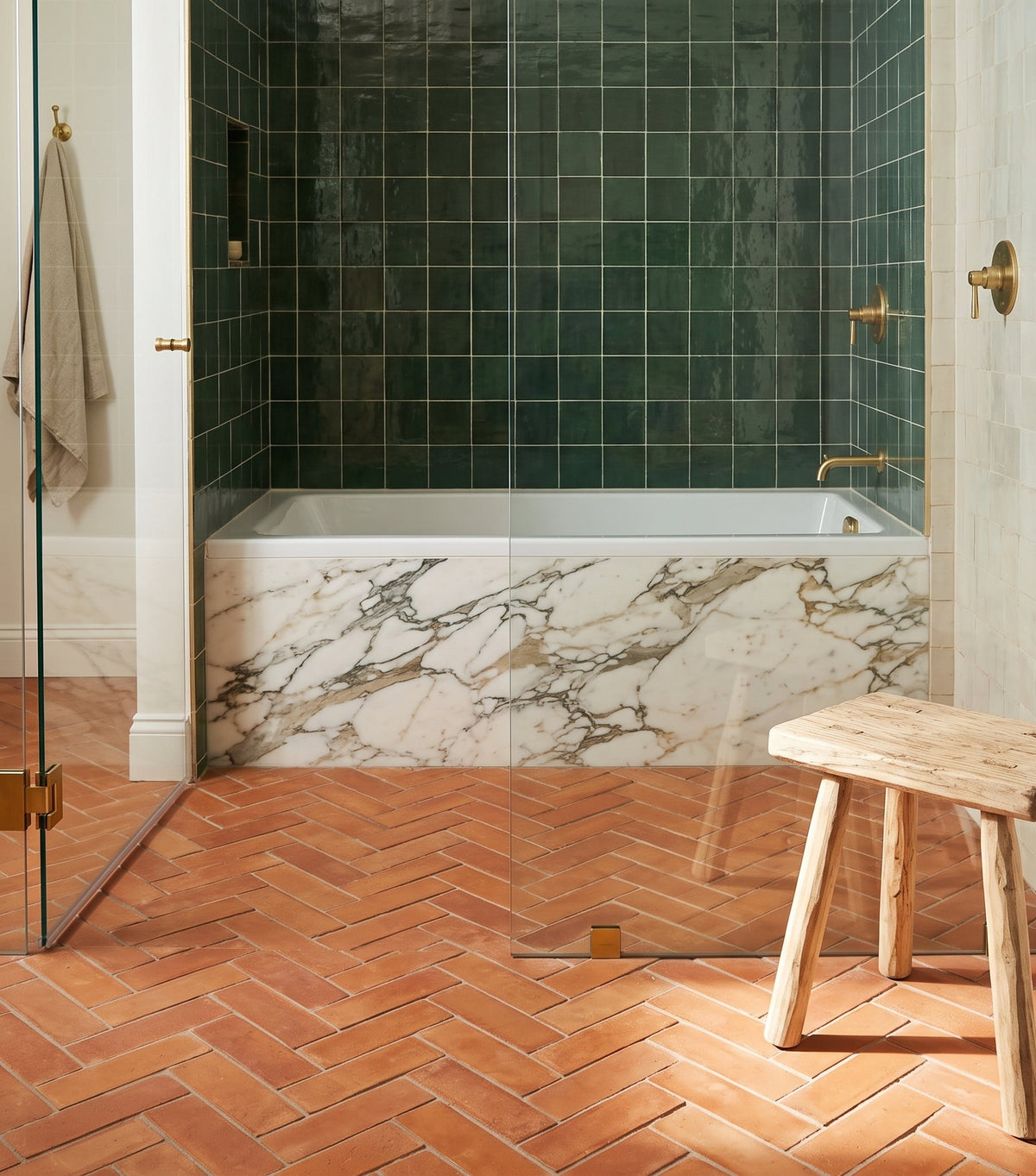 A modern bathtub with a marble front sits in a bathroom featuring dark green square tiles and terracotta herringbone flooring.