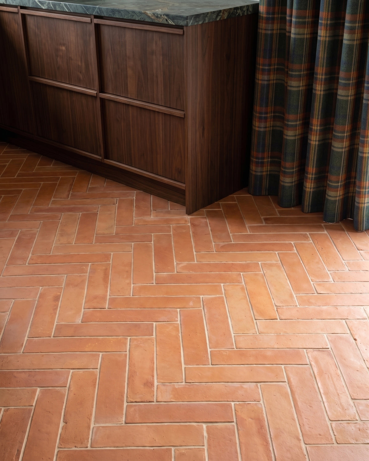Terracotta herringbone floor tiles in a kitchen setting, shown next to a dark wood cabinet with a green marble countertop and a plaid curtain.