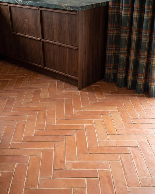Terracotta herringbone floor tiles in a kitchen setting, shown next to a dark wood cabinet with a green marble countertop and a plaid curtain.