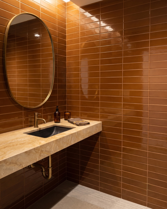 A modern bathroom corner with glossy brown rectangular wall tiles, a stone countertop, a black rectangular sink, a round wall mirror, and a small bottle and folded towel on the counter.
