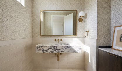 A modern bathroom vanity featuring a floating white and gray marble sink with brass fixtures, a matching rectangular mirror with a gold frame, patterned wallpaper, and light beige tile wainscoting.