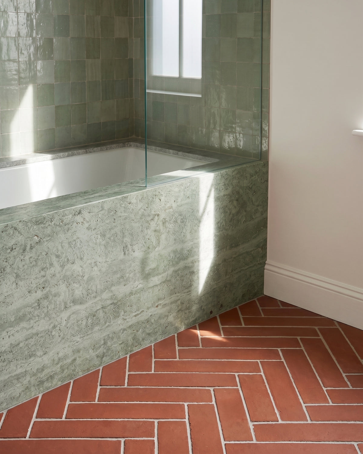Sunlight streams across a modern built-in bathtub clad in green stone, featuring glossy sage zellige wall tiles and a terracotta herringbone tile floor.
