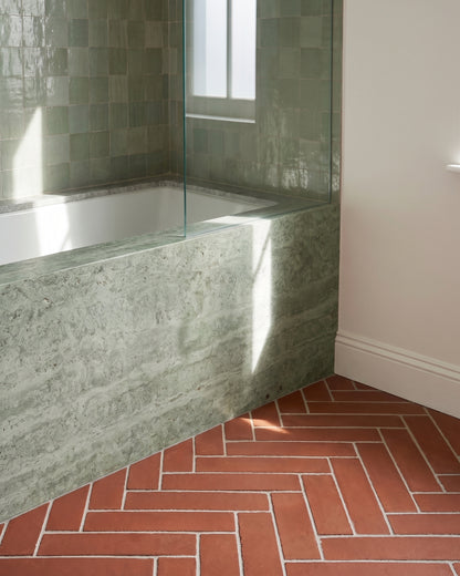 Sunlight streams across a modern built-in bathtub clad in green stone, featuring glossy sage zellige wall tiles and a terracotta herringbone tile floor.