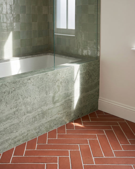 Sunlight streams across a modern built-in bathtub clad in green stone, featuring glossy sage zellige wall tiles and a terracotta herringbone tile floor.