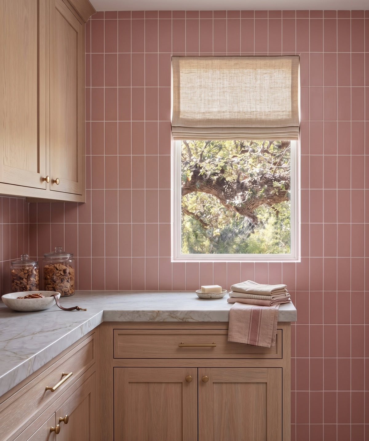 A modern kitchen features pink glossy vertical tiles, light oak cabinets, and a marble countertop accented with a stack of folded towels and a small window looking out onto green foliage.