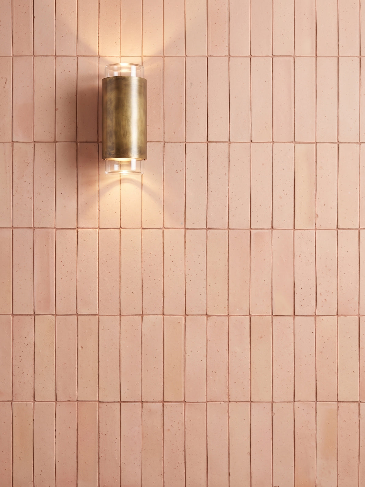 A modern cylindrical wall sconce with a brushed brass finish, mounted on a wall of small rectangular blush pink tiles and casting a warm light downward.