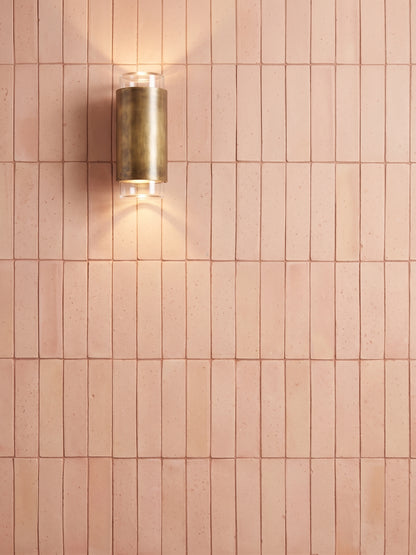 A modern cylindrical wall sconce with a brushed brass finish, mounted on a wall of small rectangular blush pink tiles and casting a warm light downward.