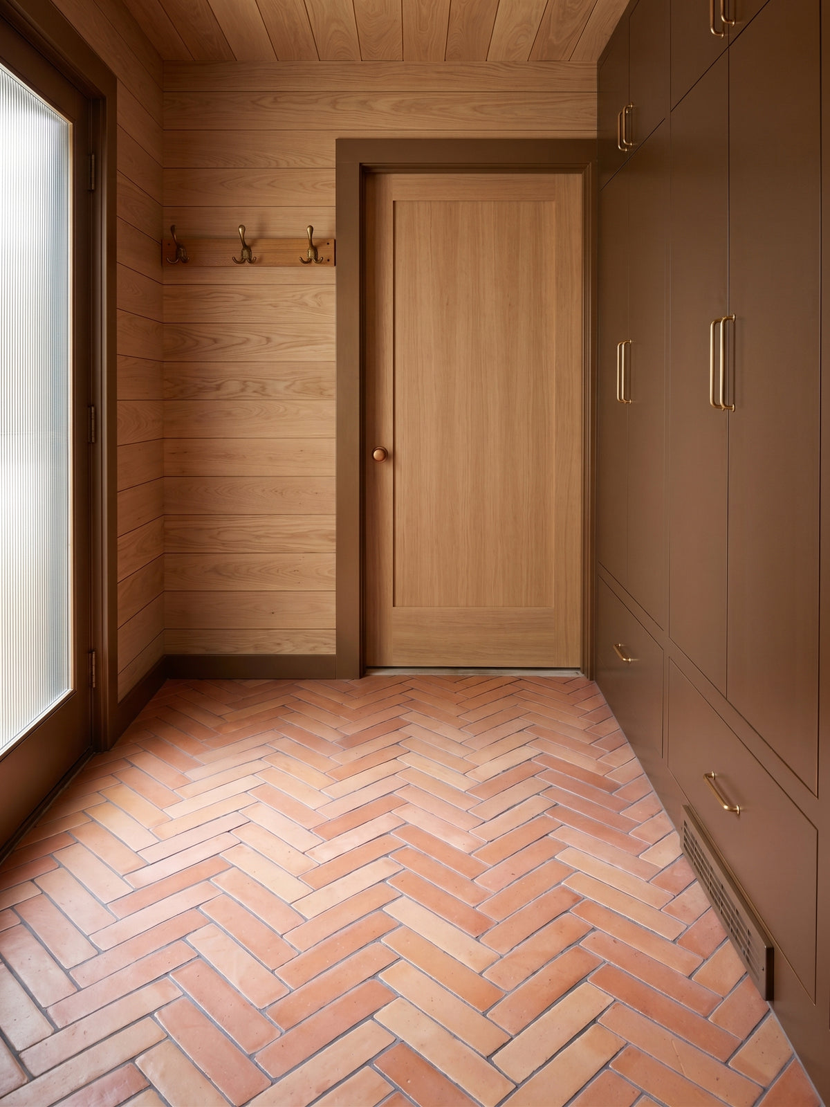 A modern interior hallway or entryway with light wood paneled walls and ceiling, a light wood door, dark brown cabinetry with brass handles, and terracotta herringbone floor tiles.