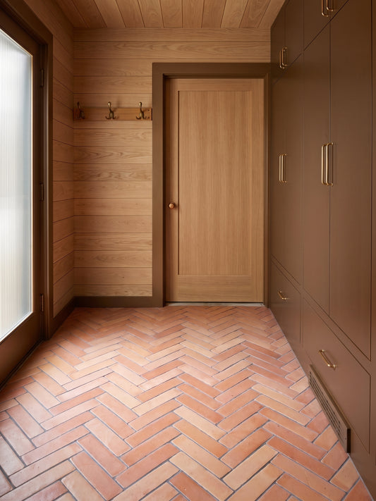 A modern interior hallway or entryway with light wood paneled walls and ceiling, a light wood door, dark brown cabinetry with brass handles, and terracotta herringbone floor tiles.