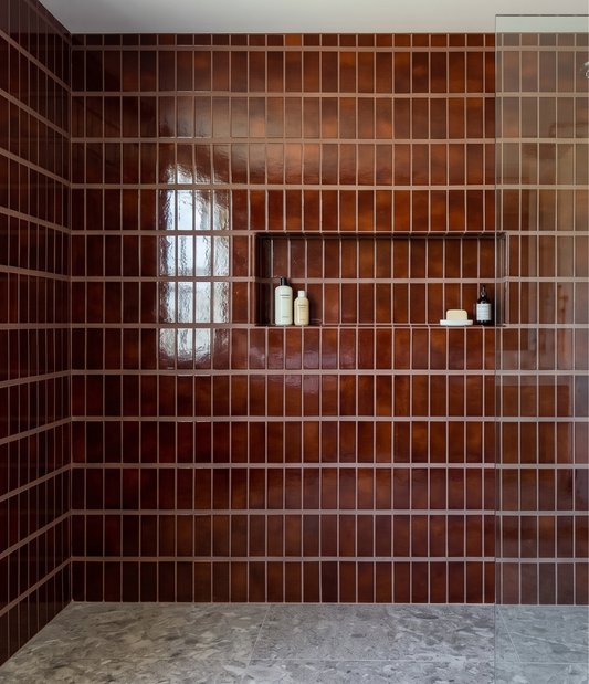 A modern shower wall featuring glossy, dark reddish-brown rectangular tiles arranged in a grid pattern, with a recessed shelf holding a few small toiletry bottles. The floor is a light gray stone tile, and a glass shower door is visible on the right side.