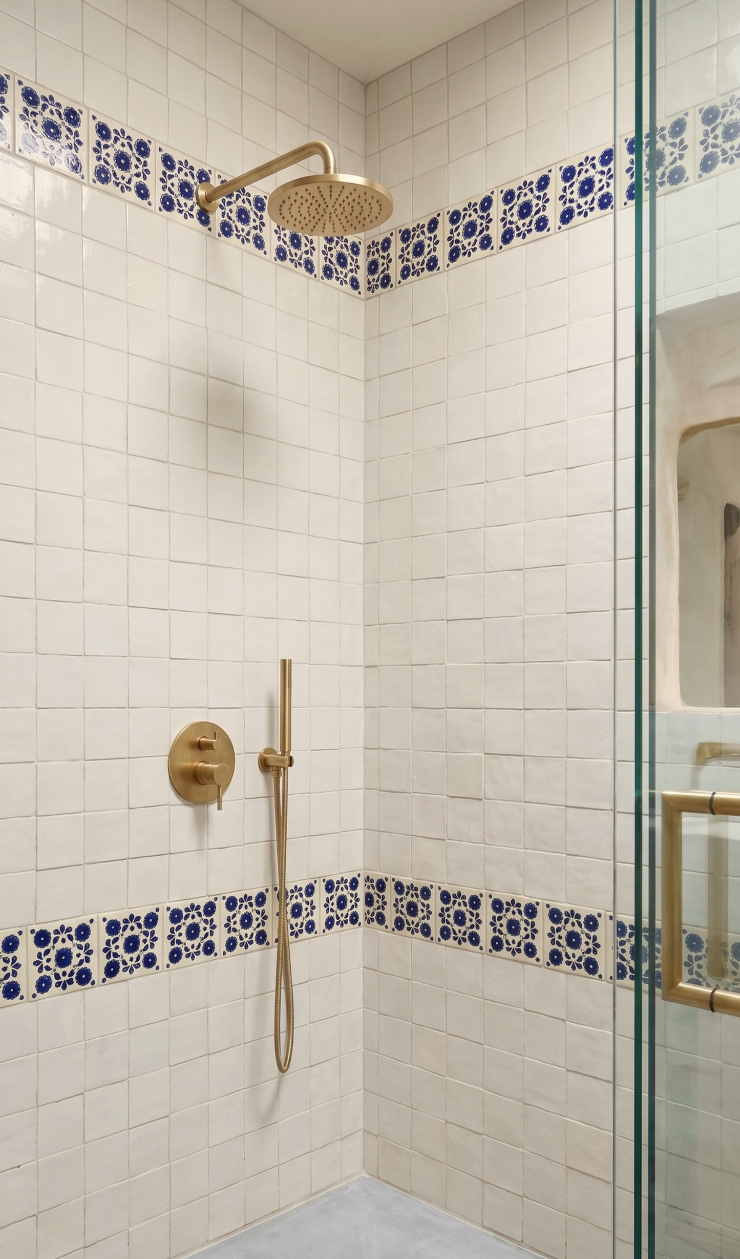 A modern walk-in shower corner with white square tiles, blue patterned tile borders, and brushed gold fixtures including a rain shower head, handheld shower, and wall controls. A glass shower door is visible on the right.
