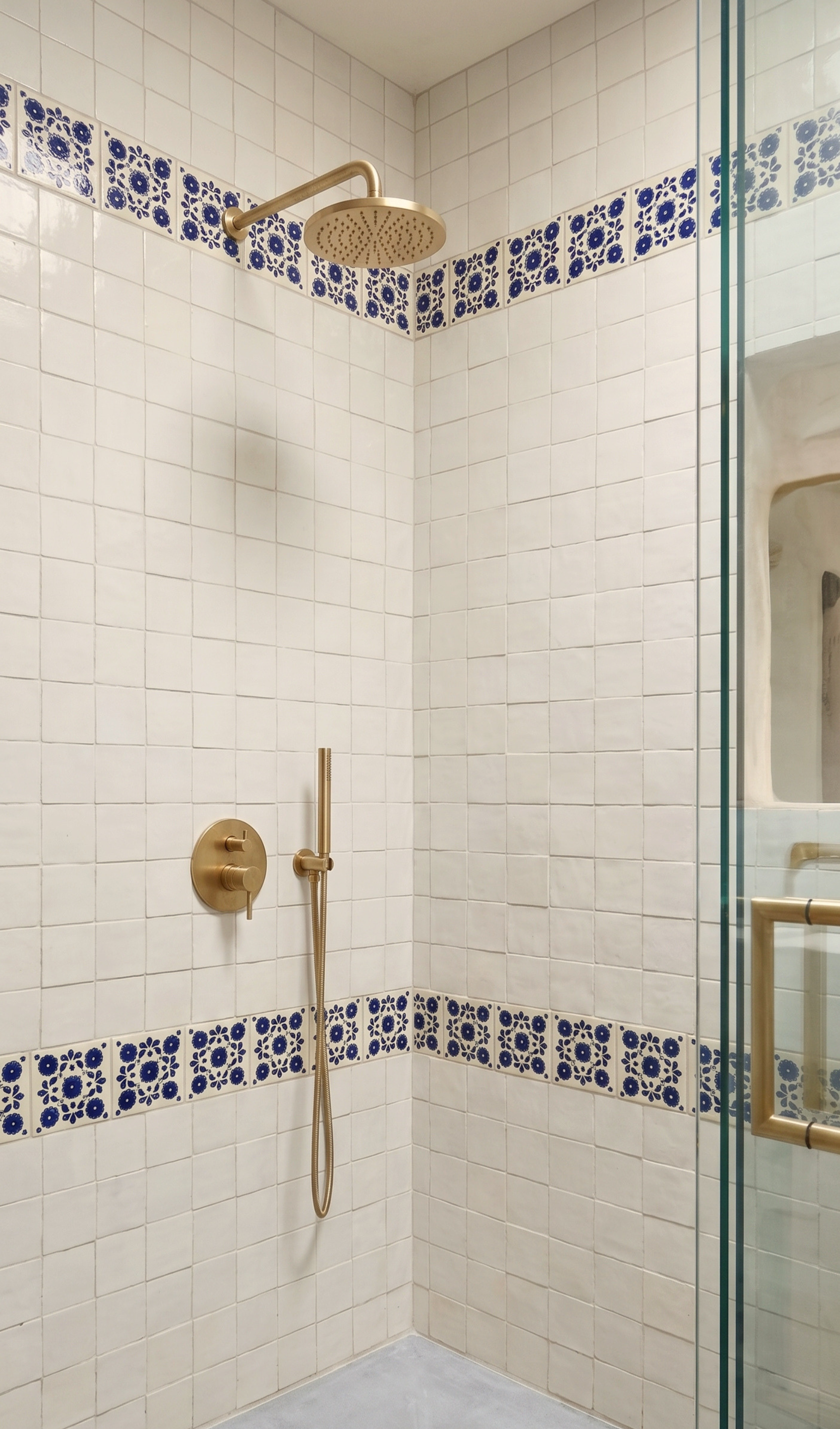 A modern walk-in shower corner with white square tiles, blue patterned tile borders, and brushed gold fixtures including a rain shower head, handheld shower, and wall controls. A glass shower door is visible on the right.
