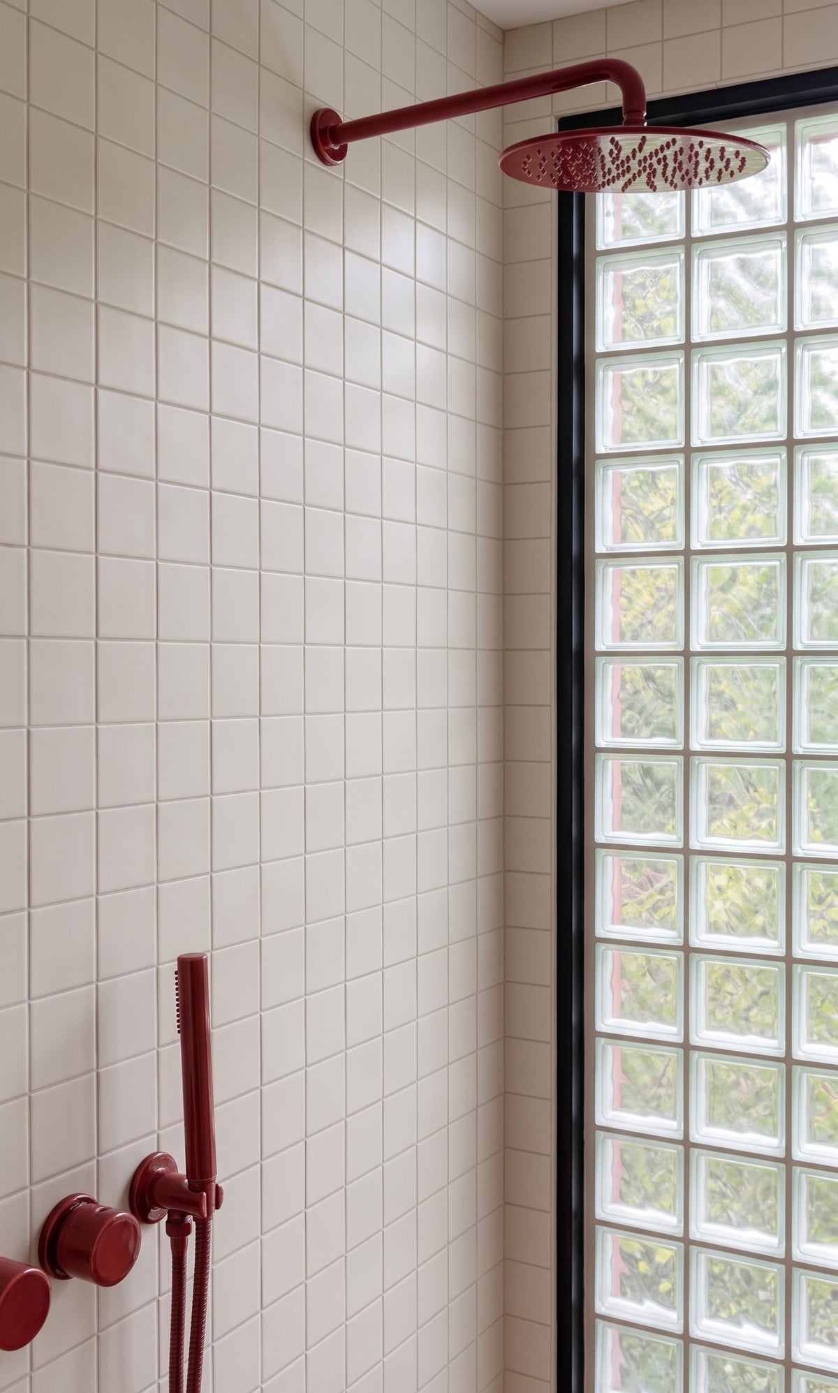 A modern bathroom corner with white square wall tiles, a dark red wall-mounted shower head and handheld shower attachment, and a glass block window with a black frame.
